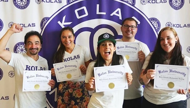group of peopl in KOLAS T-shirts holds KOLAS Univeristy certificates of completion in front of a step and repeat KOLAS logo backdrop